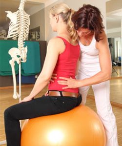 Eine Therapeutin stützt eine Patientin am Rumpf, die auf einem Gymnastikball sitzt; daneben steht ein Skelettmodell.